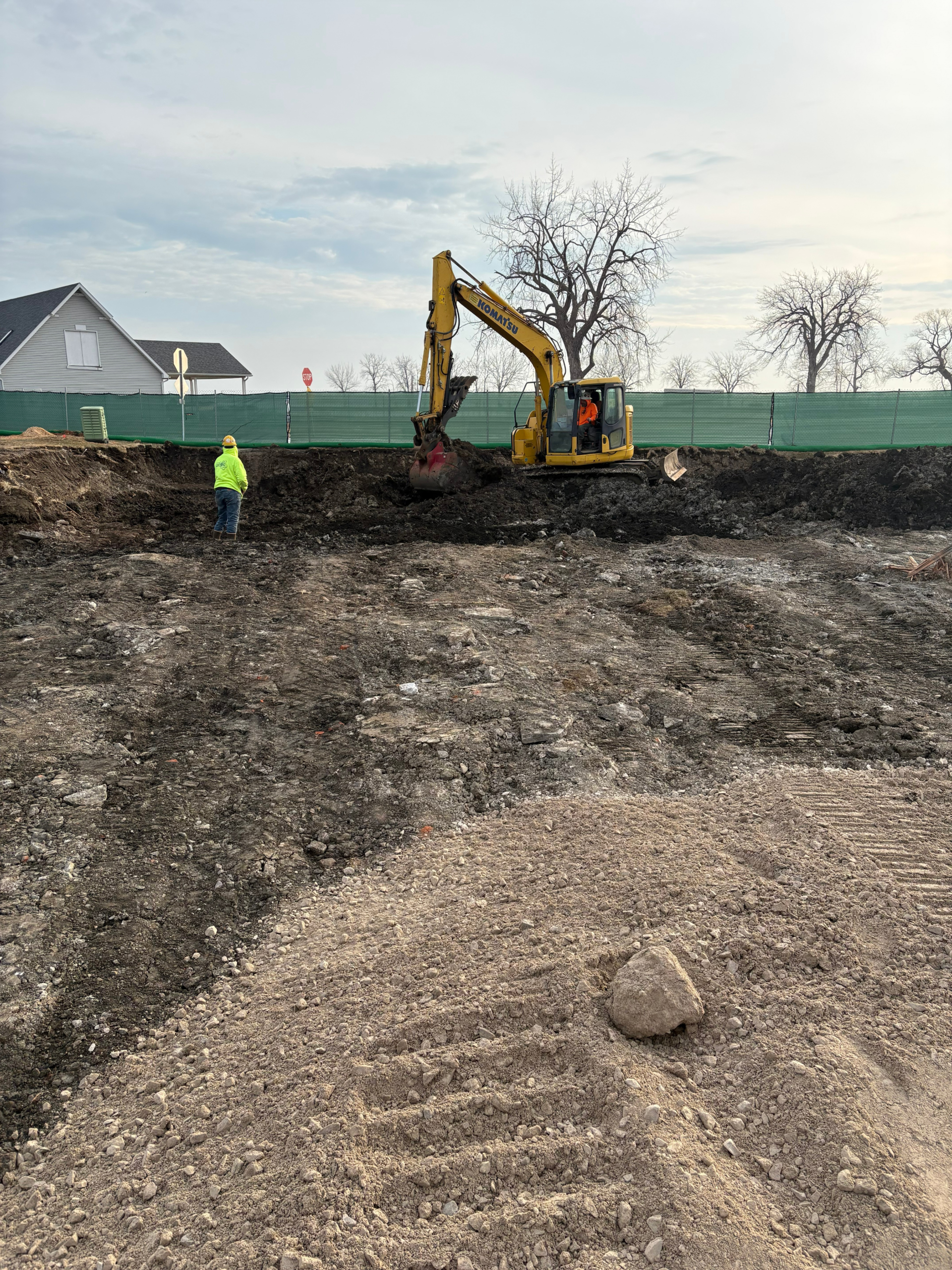 Excavator removing contaminated soil inside a fenced excavation as a worker in high-visibility PPE monitors the work at a marina remediation site