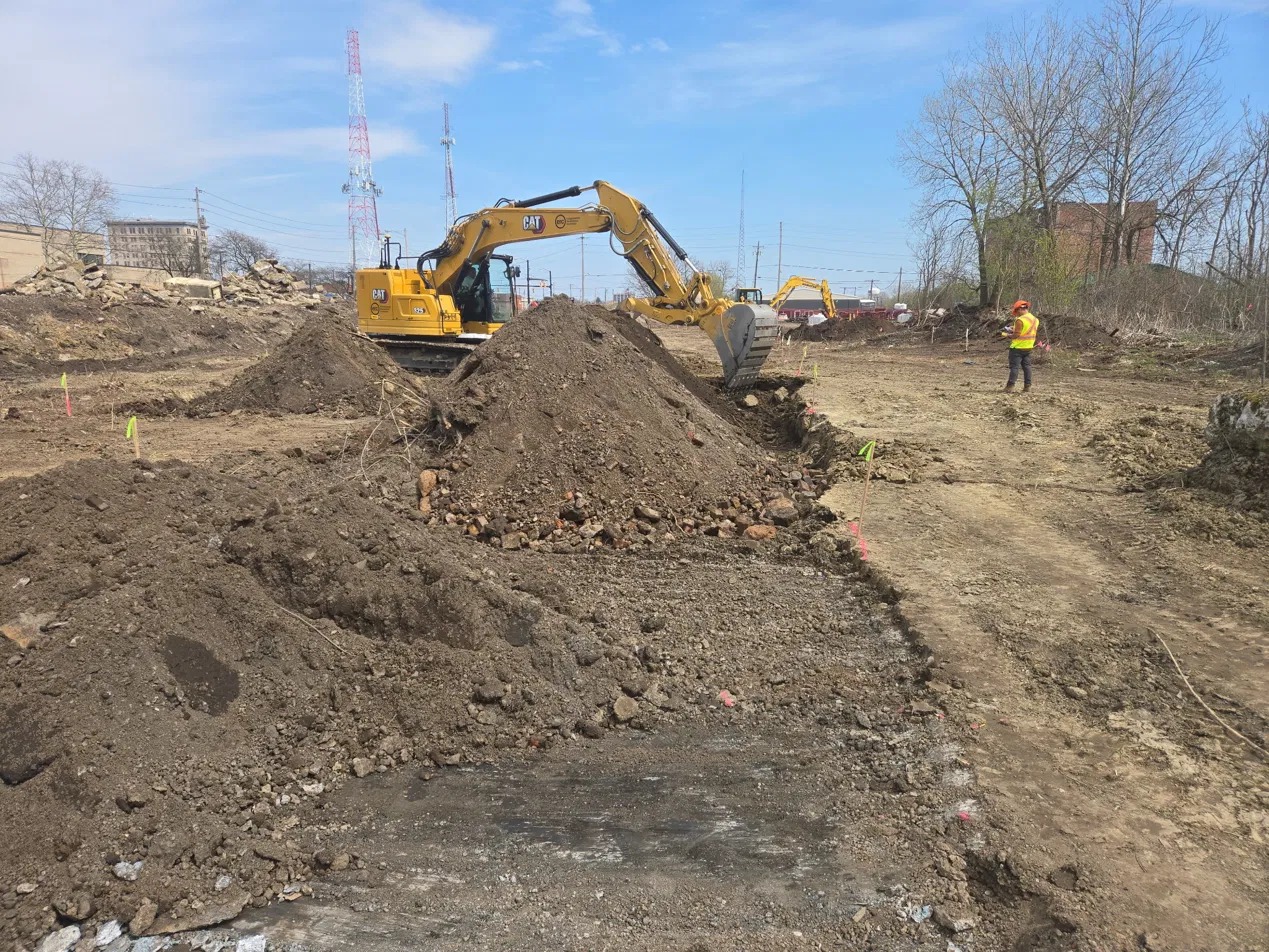 excavator beside soil stockpiles, with a worker in a hi-vis vest